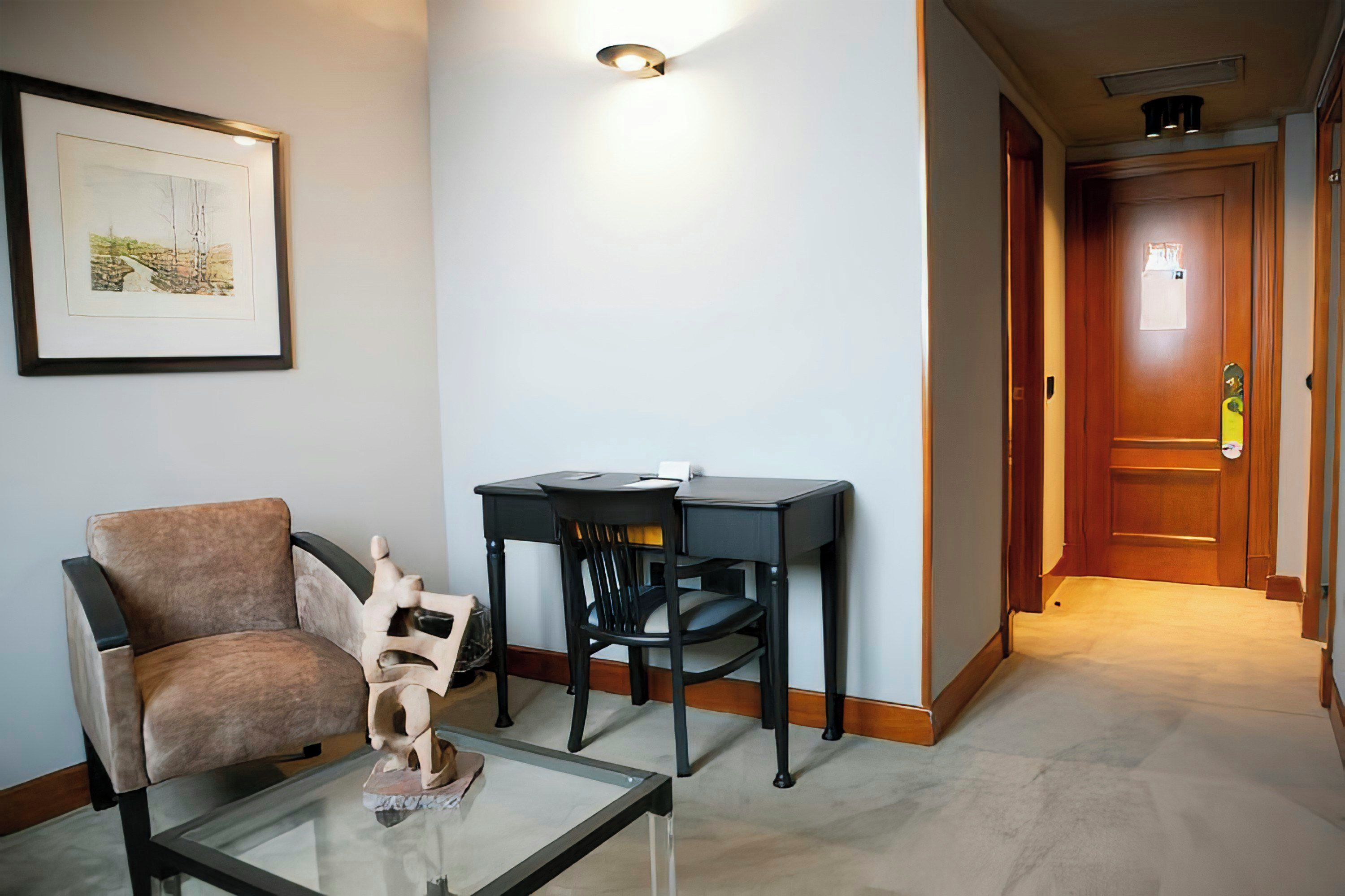 A living room with a brown armchair, a glass coffee table with a sculpture, a small black table with a chair, and a framed picture on the wall.