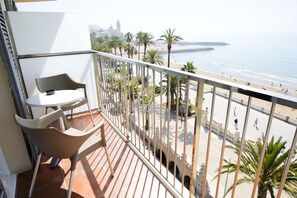 A balcony with two chairs and a table, overlooking a beach with palm trees and a distant building.
