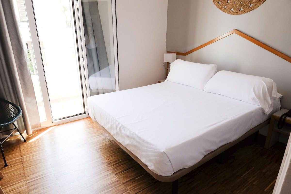 A neatly made bed with white linens in a room with wooden flooring and a large window.