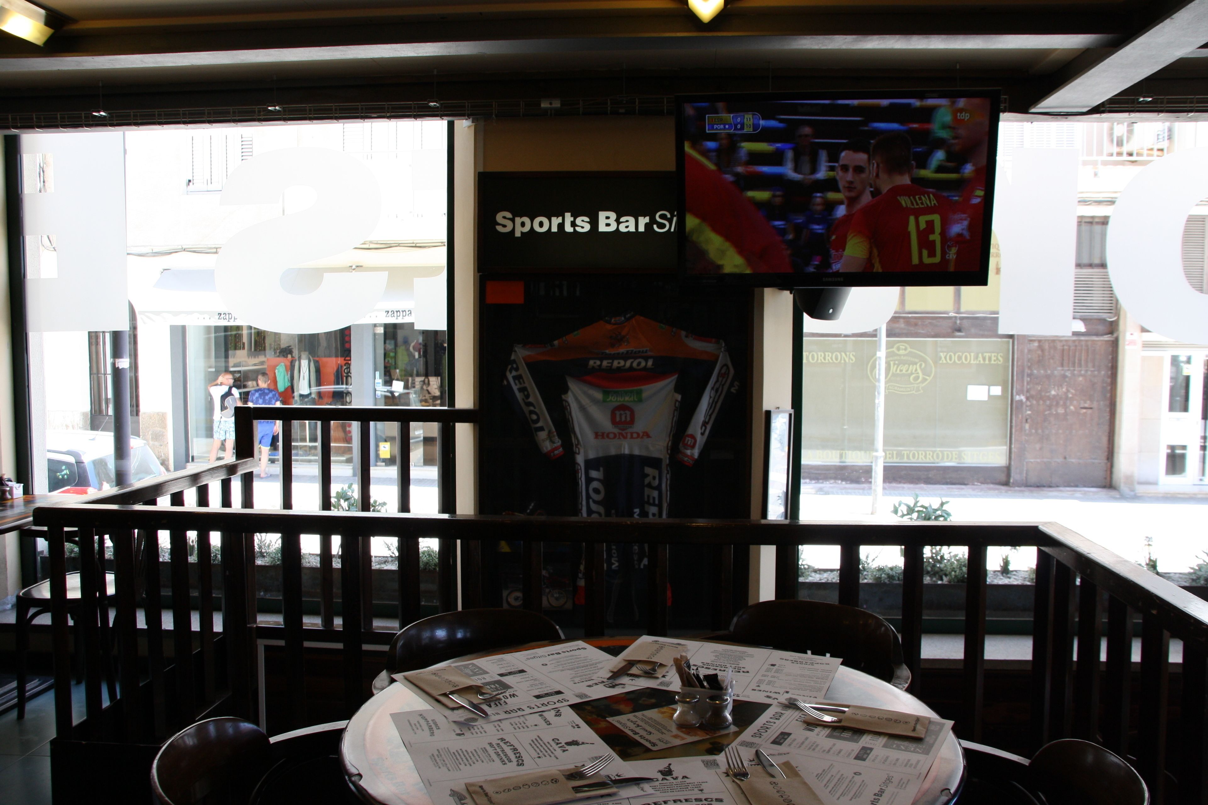 Un bar deportivo con una pantalla de televisión mostrando un partido de fútbol, un menú sobre la mesa y vistas a la calle.