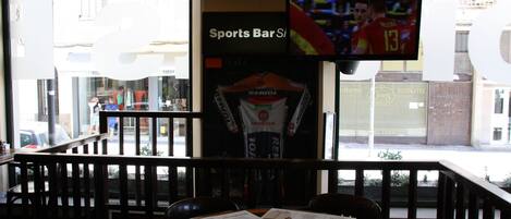 A sports bar with a TV screen displaying a soccer match, a menu on the table, and a view of the street outside.