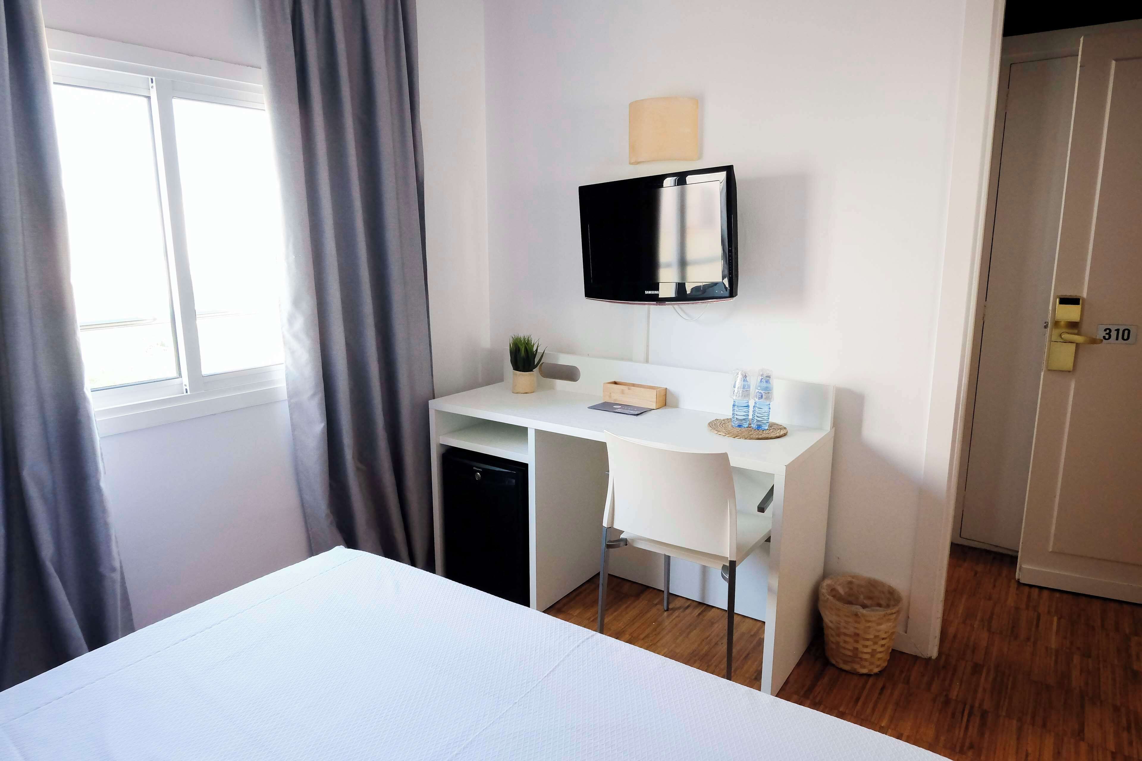 A hotel room with a bed, a desk with a computer, a TV, a small plant, and a window with curtains.