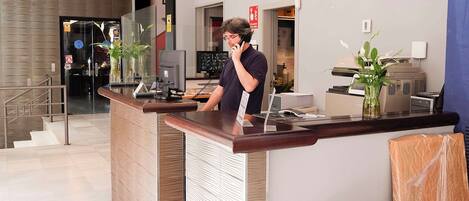 Zona de recepción de un hotel con recepcionista, mostrador de recepción, pantalla de televisión y una escalera que lleva a otra parte del hotel.