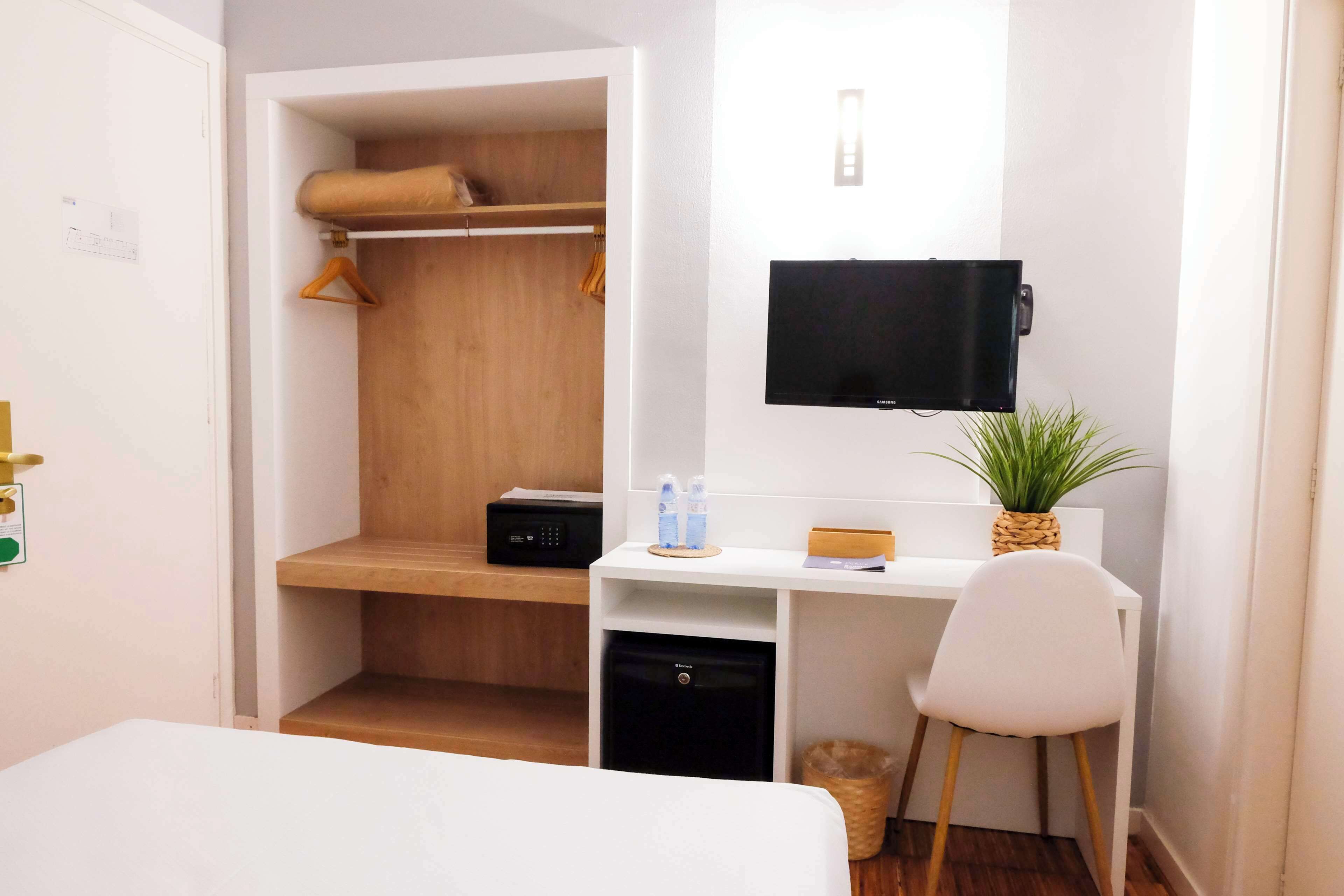 A hotel room with a bed, a desk with a computer, a TV, a small fridge, a closet, and a potted plant.