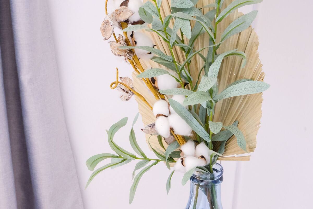 A clear glass vase with a textured design, containing dried cotton plants and green leaves, placed on a round wooden table against a plain background.