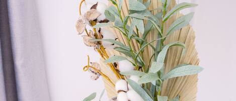 Un vase en verre transparent orné d’un motif texturé, contenant des tiges de coton séchées et des feuilles vertes, posé sur une table ronde en bois, le tout sur un fond uni.
