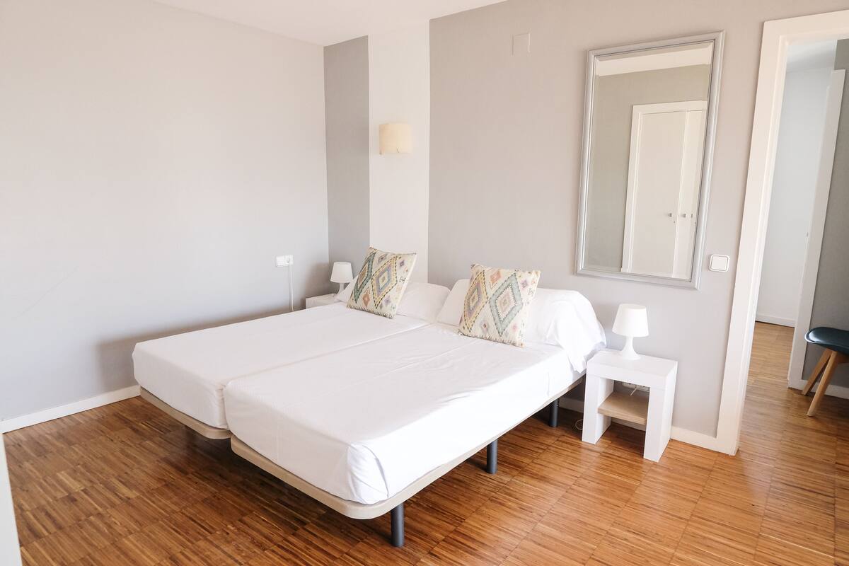 A bedroom with two beds, a bedside table with a lamp, a mirror, and a wooden floor.
