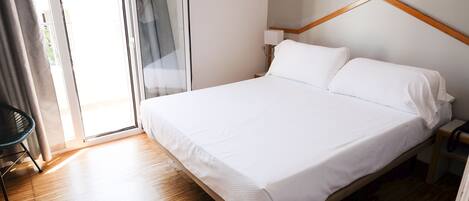 A neatly made bed with white linens in a room with wooden flooring and a large window.