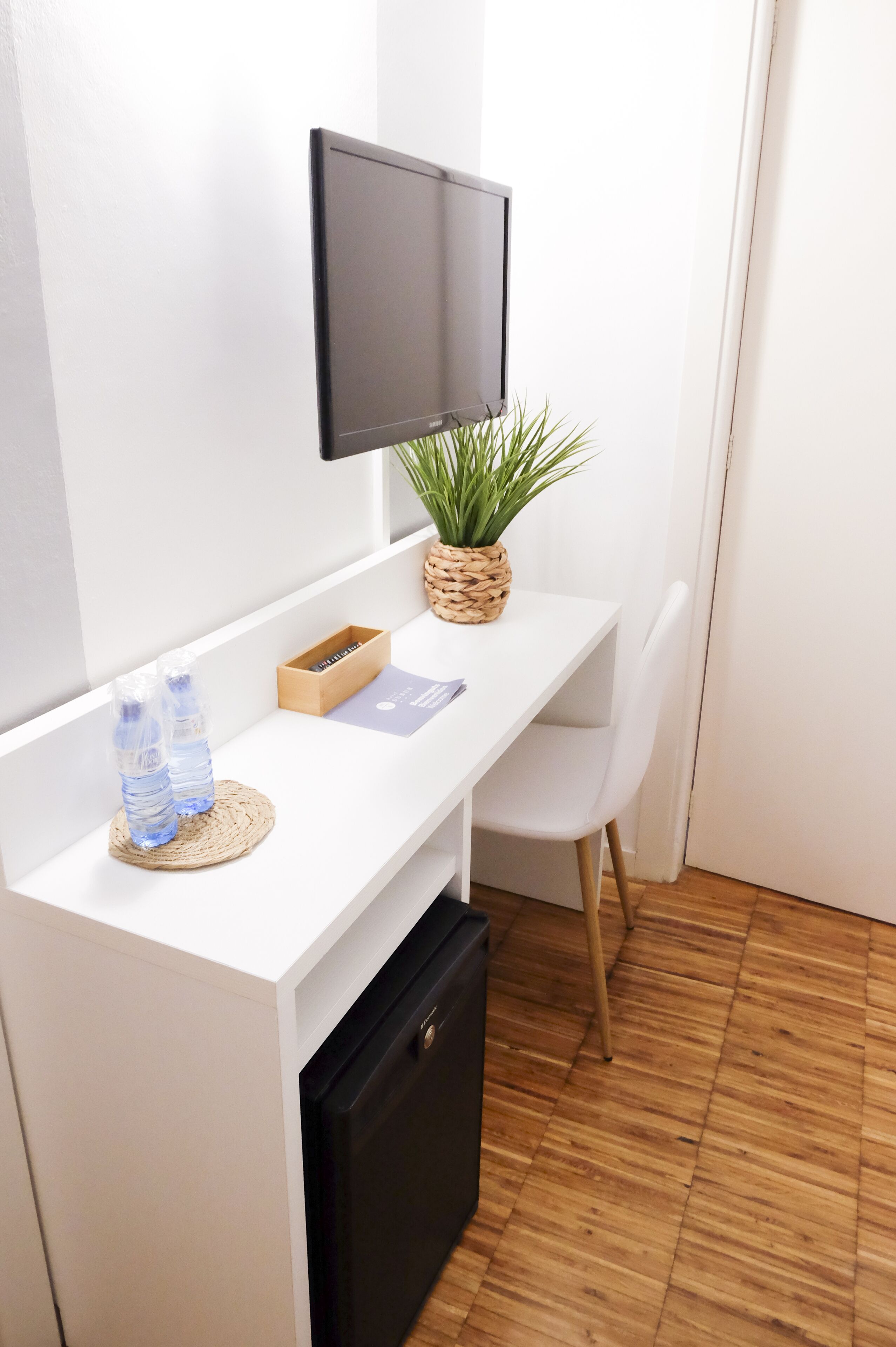 A small, white desk with a flat-screen TV mounted above it, a woven basket with a plant, a water bottle, and a box of tissues.