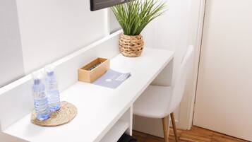 A small, white desk with a flat-screen TV mounted above it, a woven basket with a plant, a water bottle, and a box of tissues.