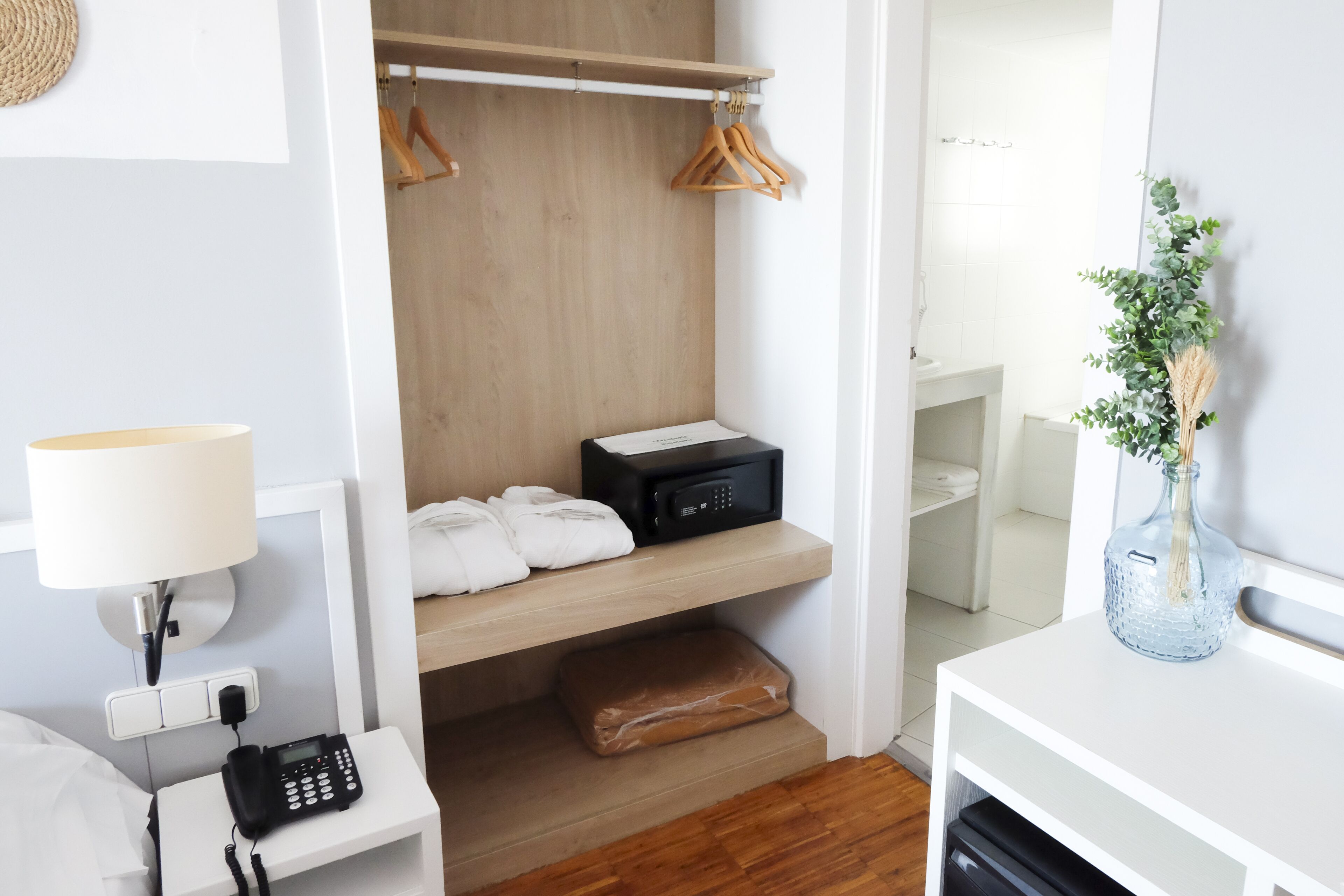 A hotel room with a wooden closet, a bedside phone, a lamp, a small table with a vase, and a bathroom visible through an open door.