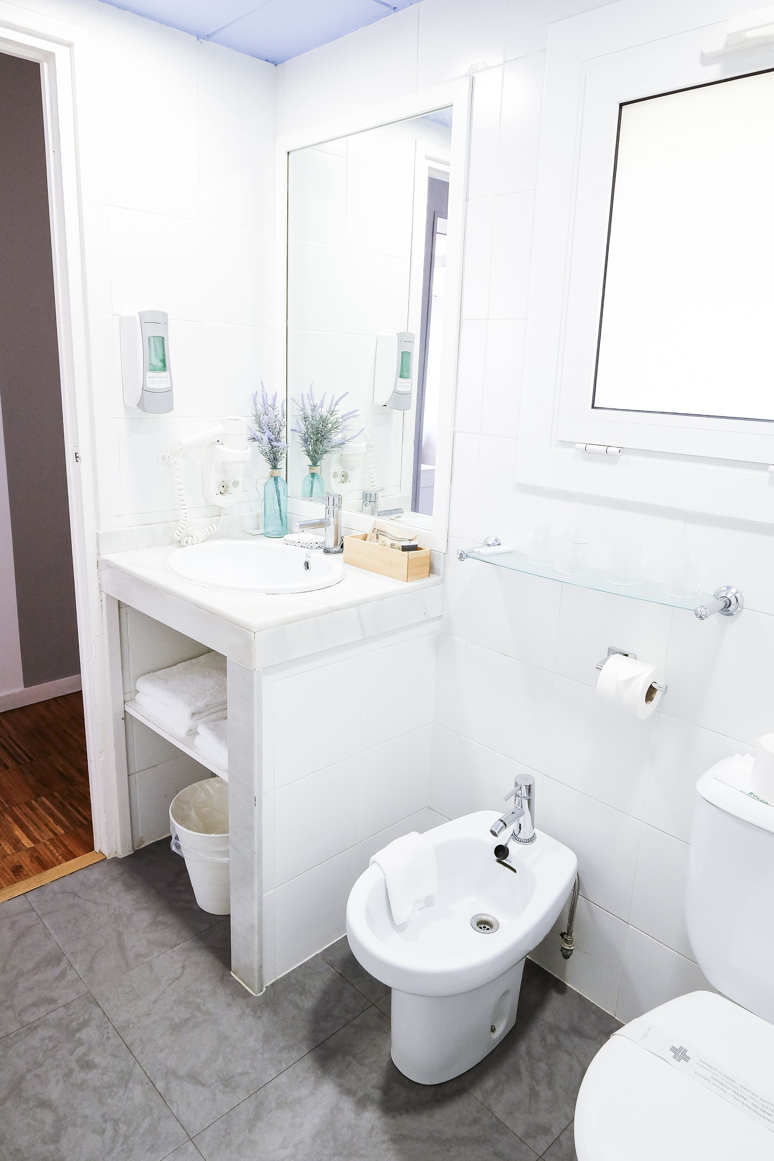 Une salle de bain moderne avec un lavabo blanc, un bidet et des toilettes. Il y a une étagère en verre au-dessus du lavabo et un miroir sur le mur.