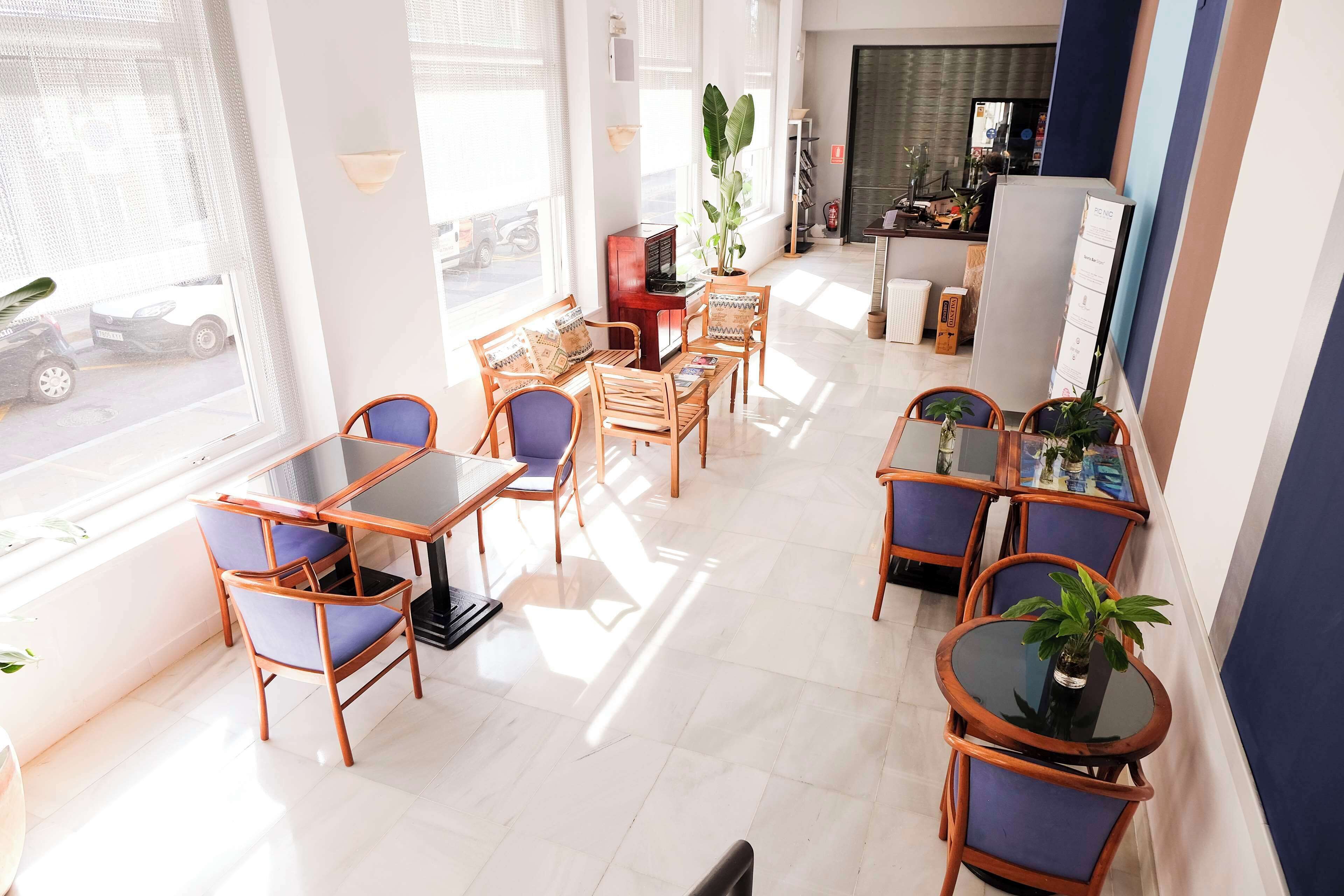 A well-lit dining area with wooden tables and chairs, large windows, and potted plants.