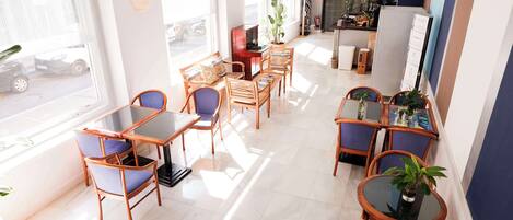 A well-lit dining area with wooden tables and chairs, large windows, and potted plants.
