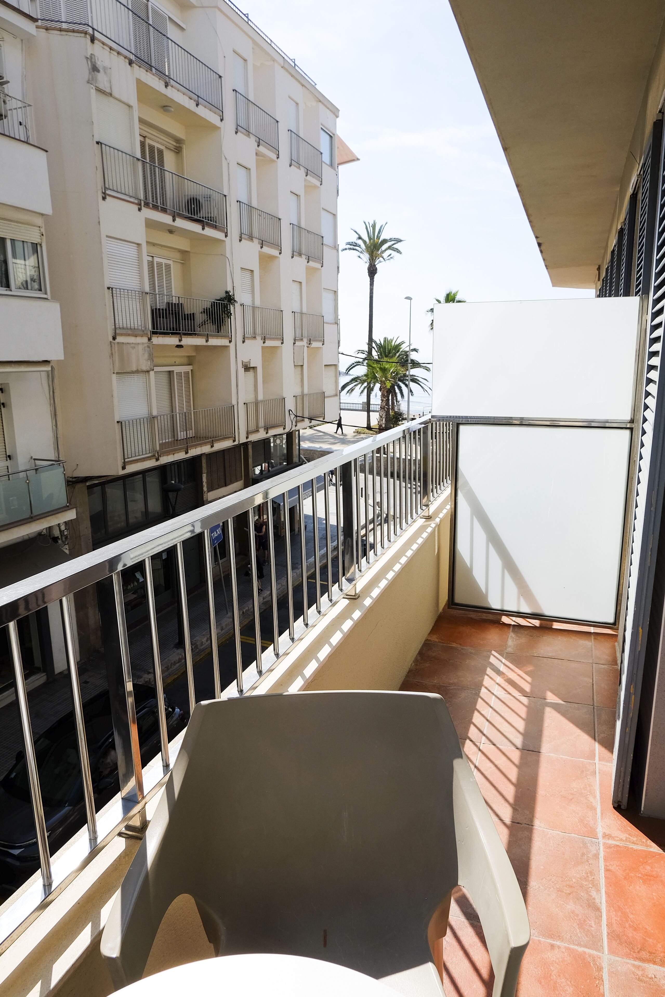 Un balcón con una silla blanca, un refrigerador blanco y vistas de un edificio de varios pisos con balcones y ventanas.