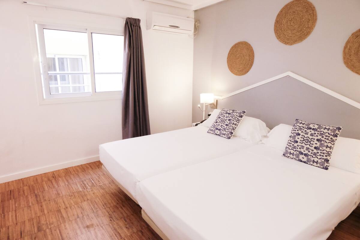 A double bed with white bedding and patterned pillows, a window with curtains, and a wall with woven decorations.