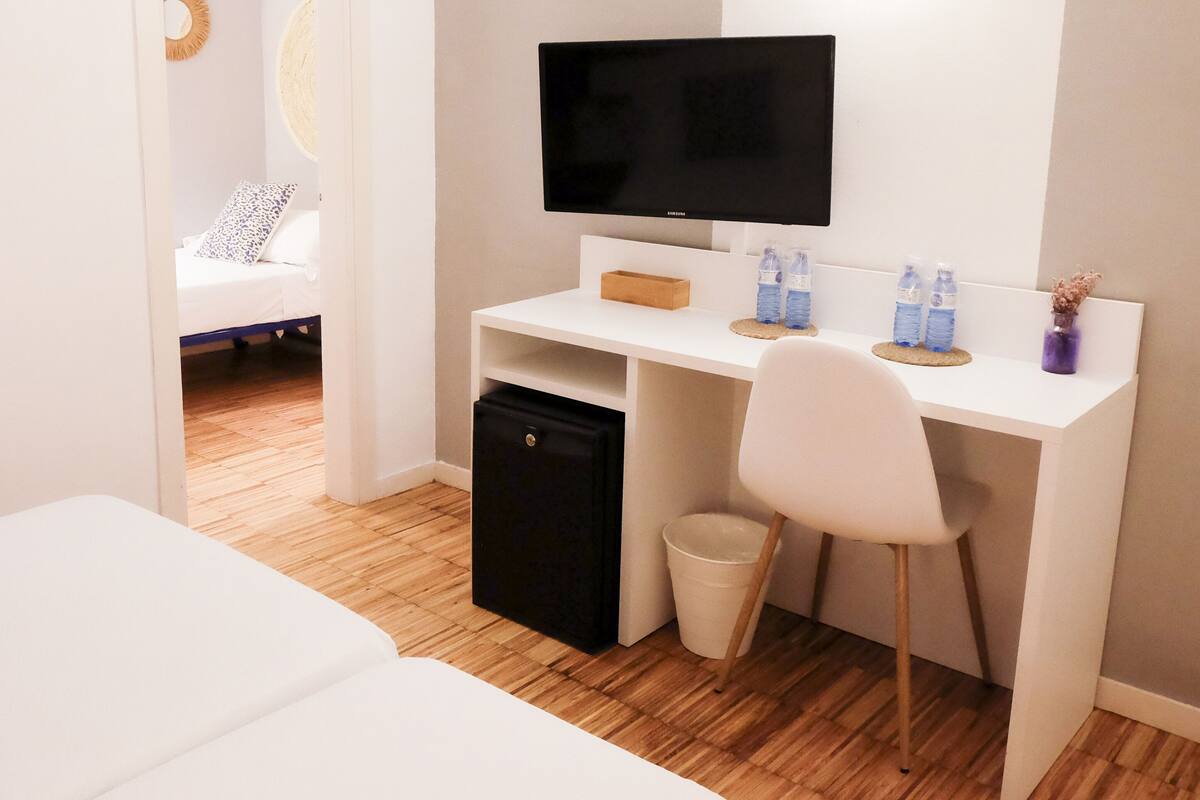 A hotel room with a bed, a desk with a computer, a chair, a small fridge, and a trash bin.