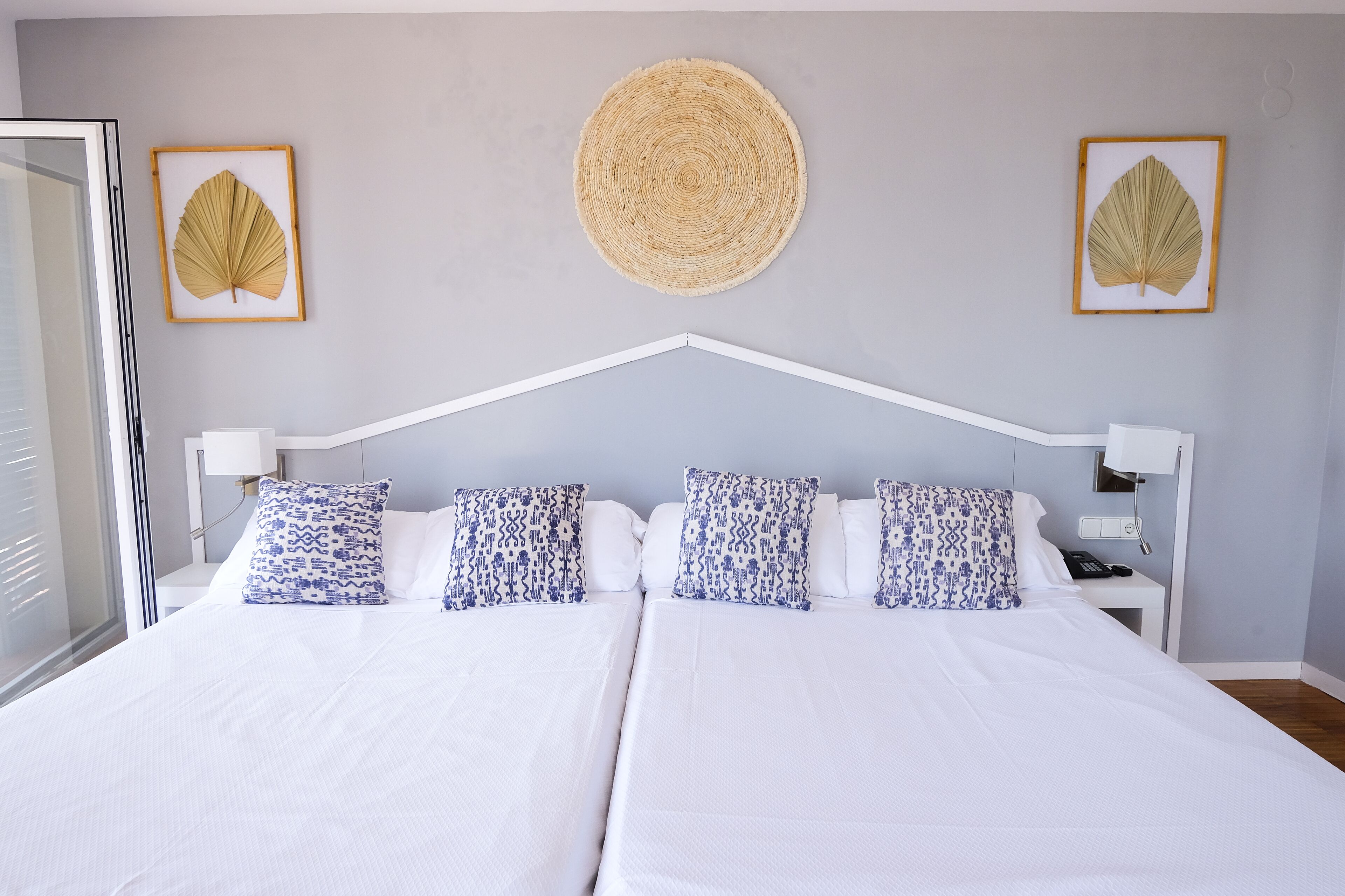 A double bed with white bedding and blue patterned pillows, a woven wall hanging, and framed leaf artwork.