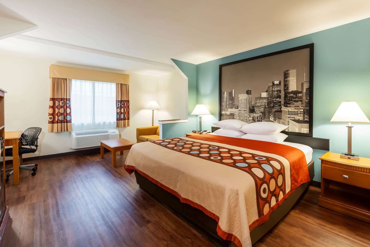 A hotel room with a large bed, two bedside tables with lamps, a desk with a chair, a window with curtains, and a cityscape painting on the wall.