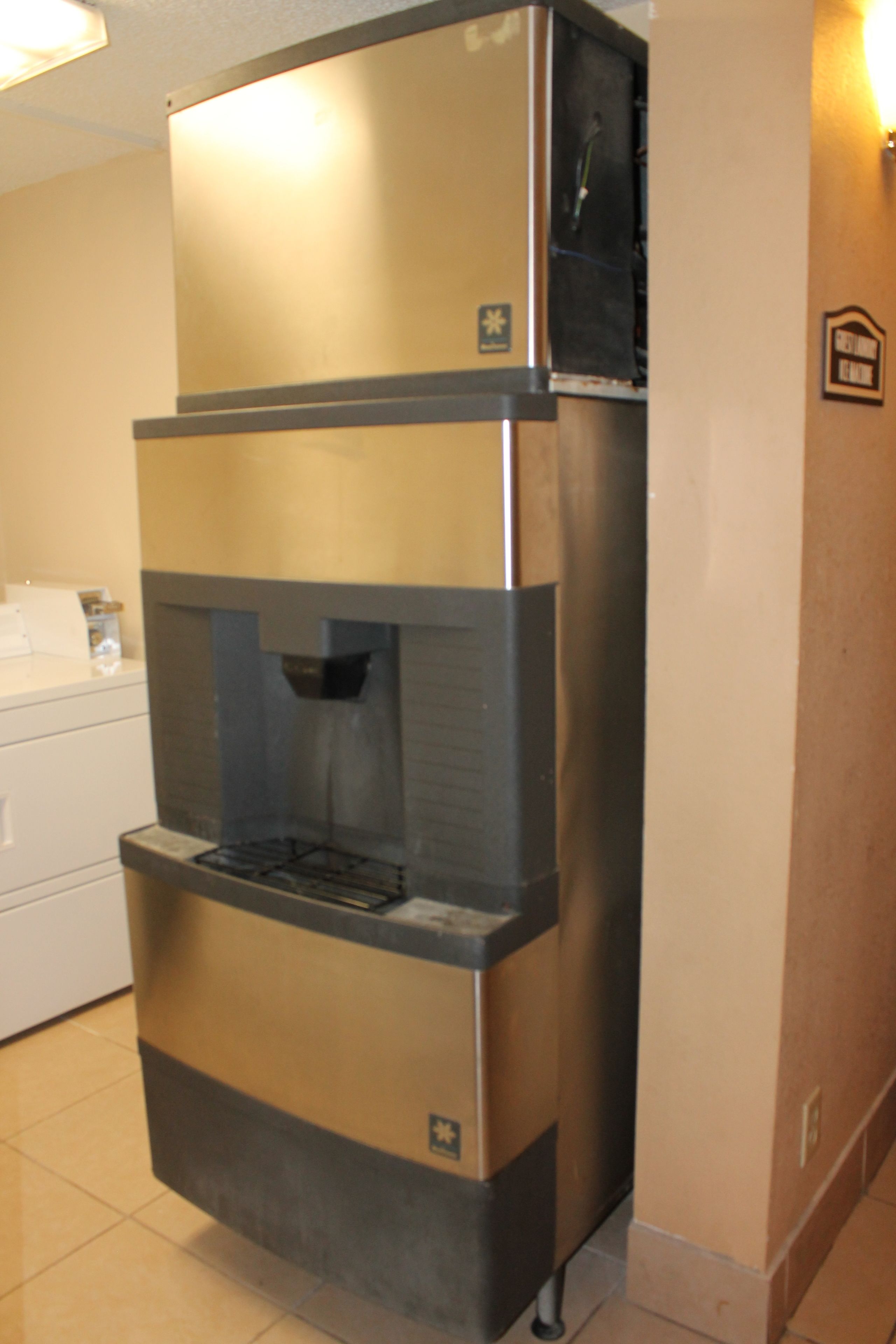A stainless steel coffee machine with a water dispenser, located in a kitchen area with a white countertop and a wall-mounted sign.