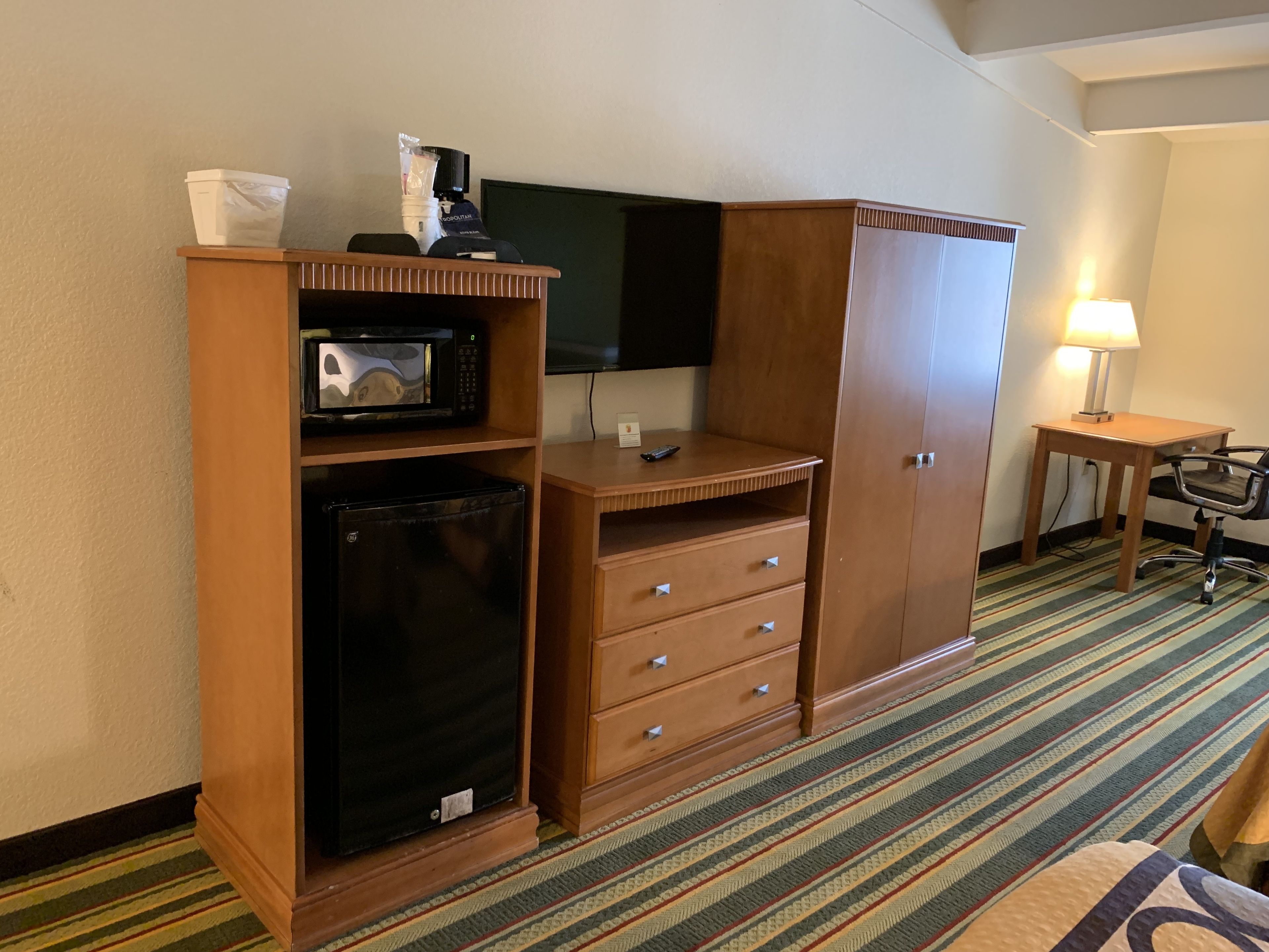 Habitación de hotel con kitchenette, televisor de pantalla plana, escritorio con silla y alfombra a rayas.