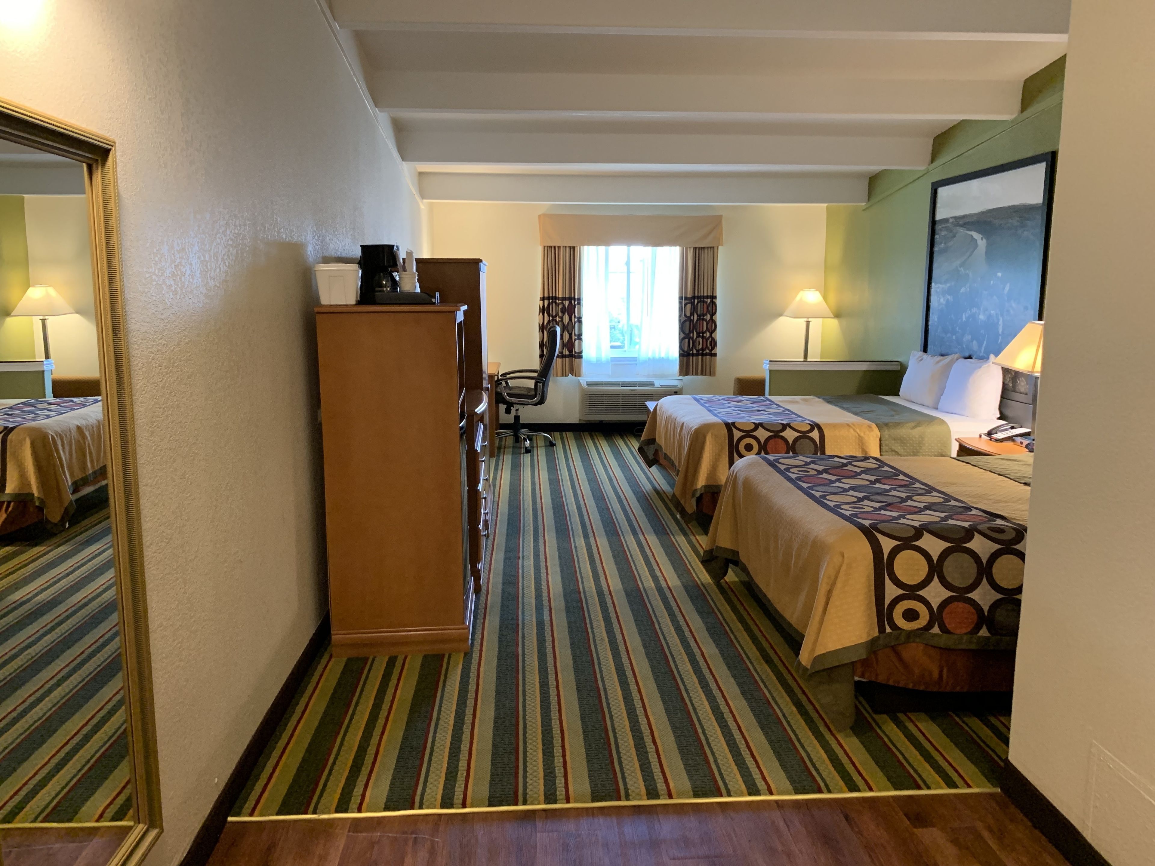 A hotel room with two beds, a desk, a chair, a mirror, and a window with curtains.