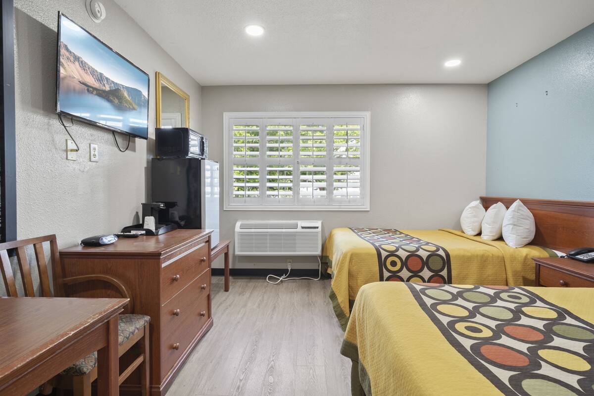 A hotel room with two beds, a desk, a television, a window with blinds, and an air conditioning unit.
