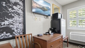 A hotel room with a wooden dresser, a television mounted on the wall, a large window with blinds, and a painting on the wall. - Travelers Inn Medford (Medford)