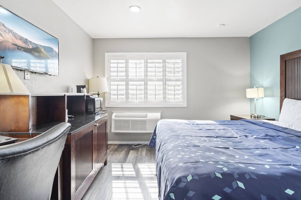 A hotel room with a bed, a desk, a chair, a window with blinds, a wall-mounted TV, and a bedside lamp.