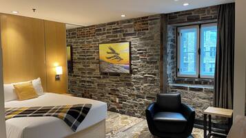 Quarto de hotel com uma cama, uma poltrona de couro preto, uma mesinha, parede de pedra e janela com vista para edifícios.