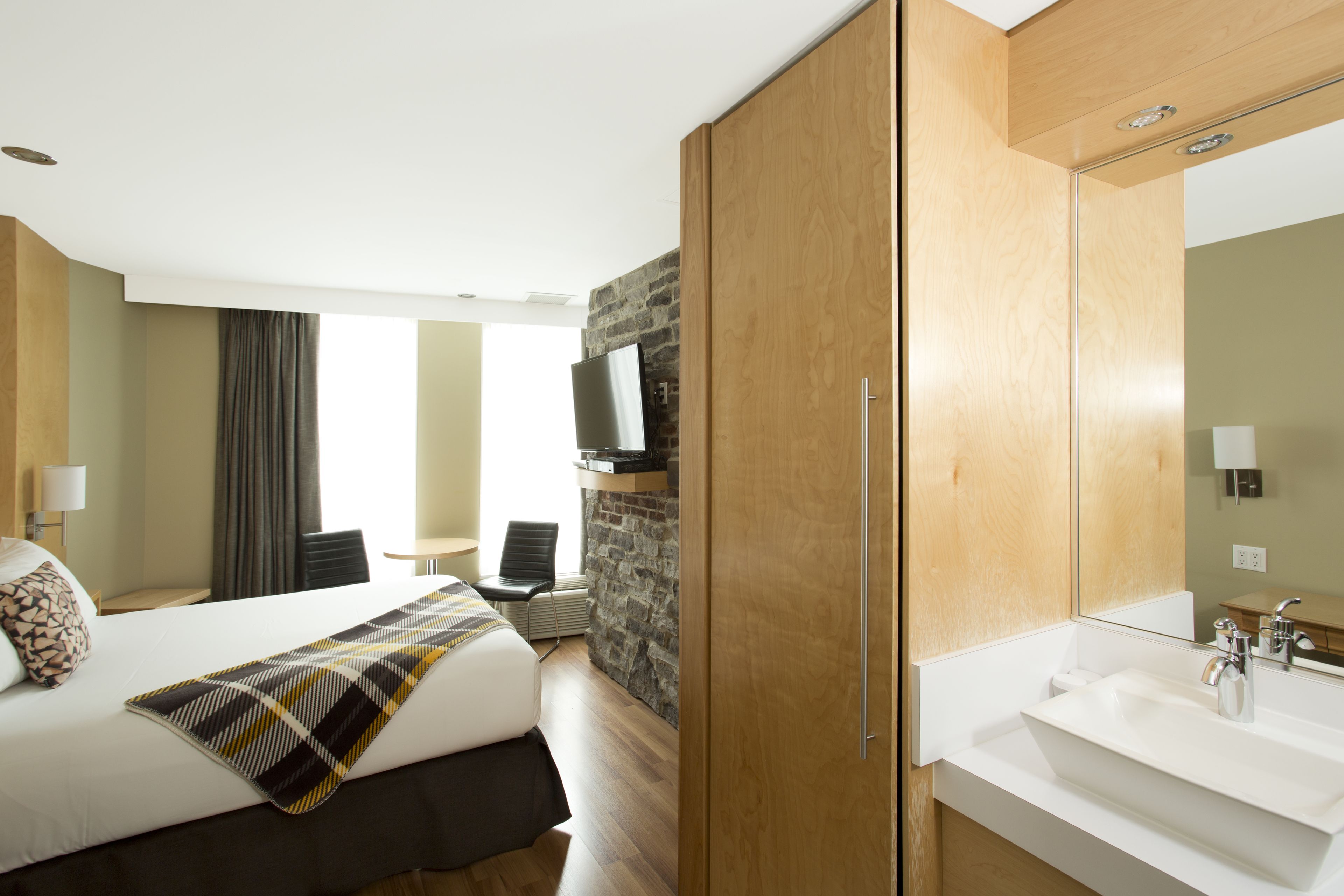 Quarto de hotel com cama, guarda-roupa de madeira, banheiro com pia e espelho, e parede de destaque em pedra.
