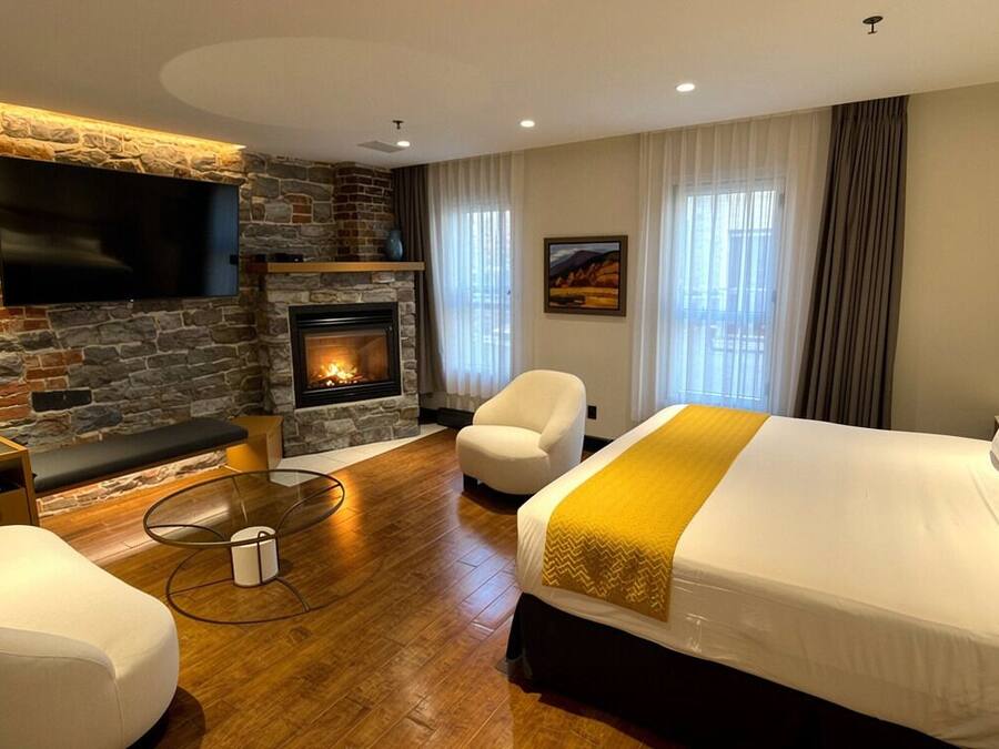 A modern hotel room with a stone wall, a fireplace, a large bed, a sofa, a TV, and a view of the outdoors.