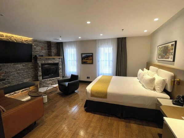 Habitación de hotel con chimenea de piedra, una cama con ropa blanca, una silla moderna y un televisor de pantalla plana montado en la pared.