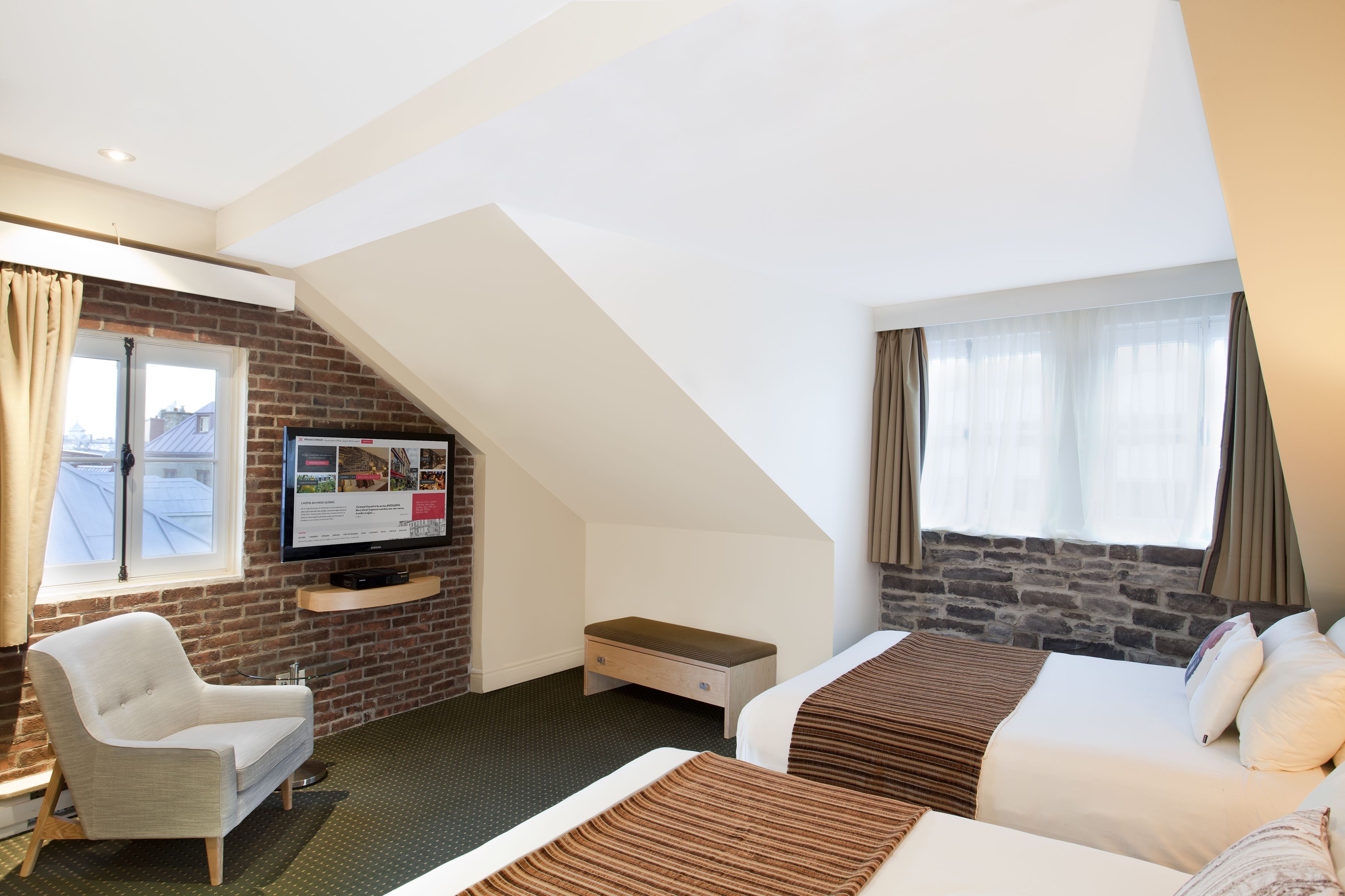 Quarto de hotel com parede de tijolos, televisão montada na parede, uma escrivaninha, uma cadeira e duas camas.