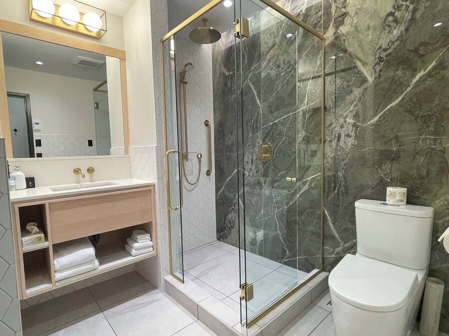 Un baño moderno con ducha acristalada, pared de mármol, inodoro blanco y tocador con espejo e iluminación.
