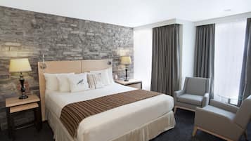 Une chambre d’hôtel avec un mur en pierre, un grand lit avec une literie blanche, deux tables de chevet avec des lampes, un fauteuil gris et une vue sur la ville à travers de grandes fenêtres.
