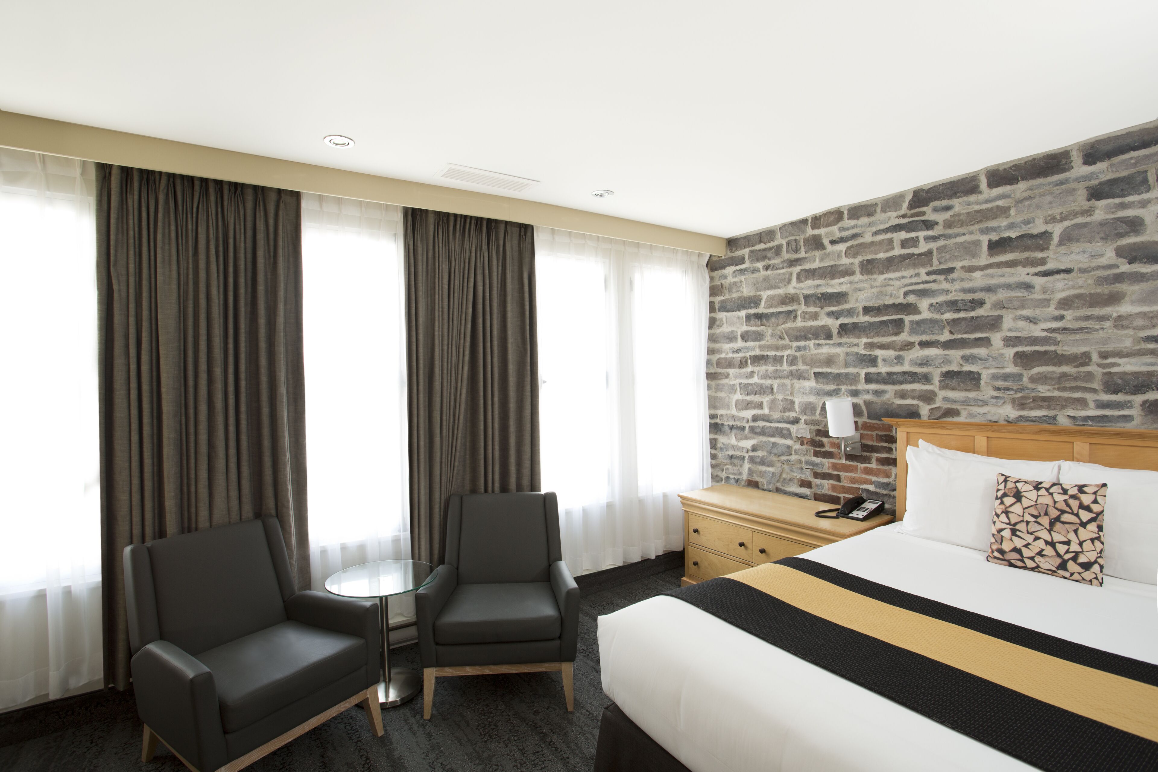 A hotel room with a large bed, two armchairs, a small table, a nightstand with a lamp, and a stone wall.