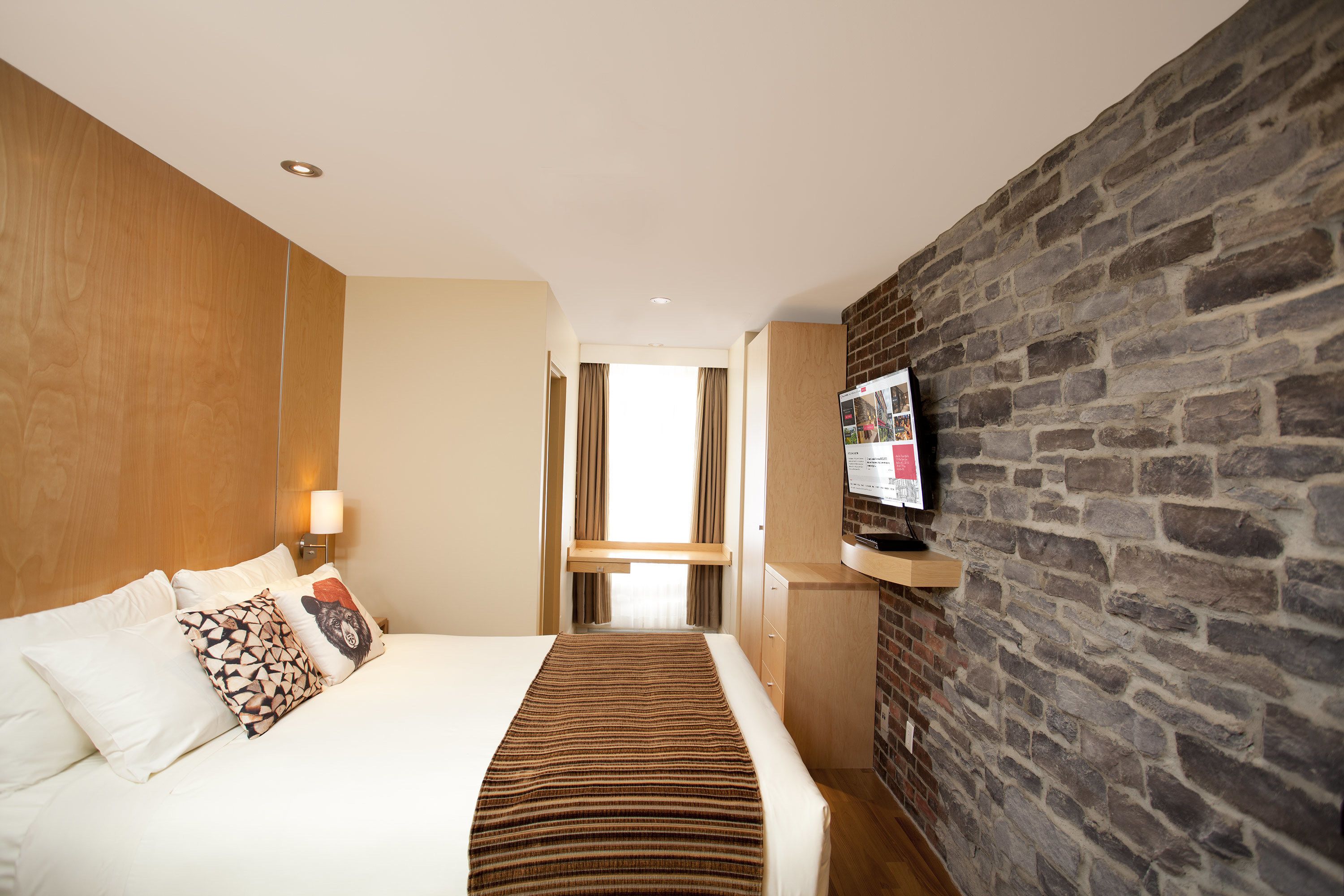Une chambre d’hôtel avec un lit, un téléviseur fixé au mur en briques et une fenêtre avec des rideaux.