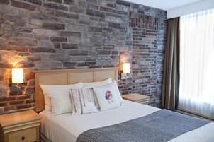 A hotel room with a brick wall, a bed with white bedding, two bedside lamps, and a window with curtains.