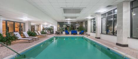 An indoor swimming pool with lounge chairs and tables, surrounded by large windows and indoor plants.