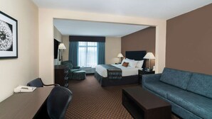 A hotel room with a bed, a sofa, a desk, and a chair. There is a window with curtains, a lamp, and a framed picture on the wall. - Wingate by Wyndham Lexington (Lexington)