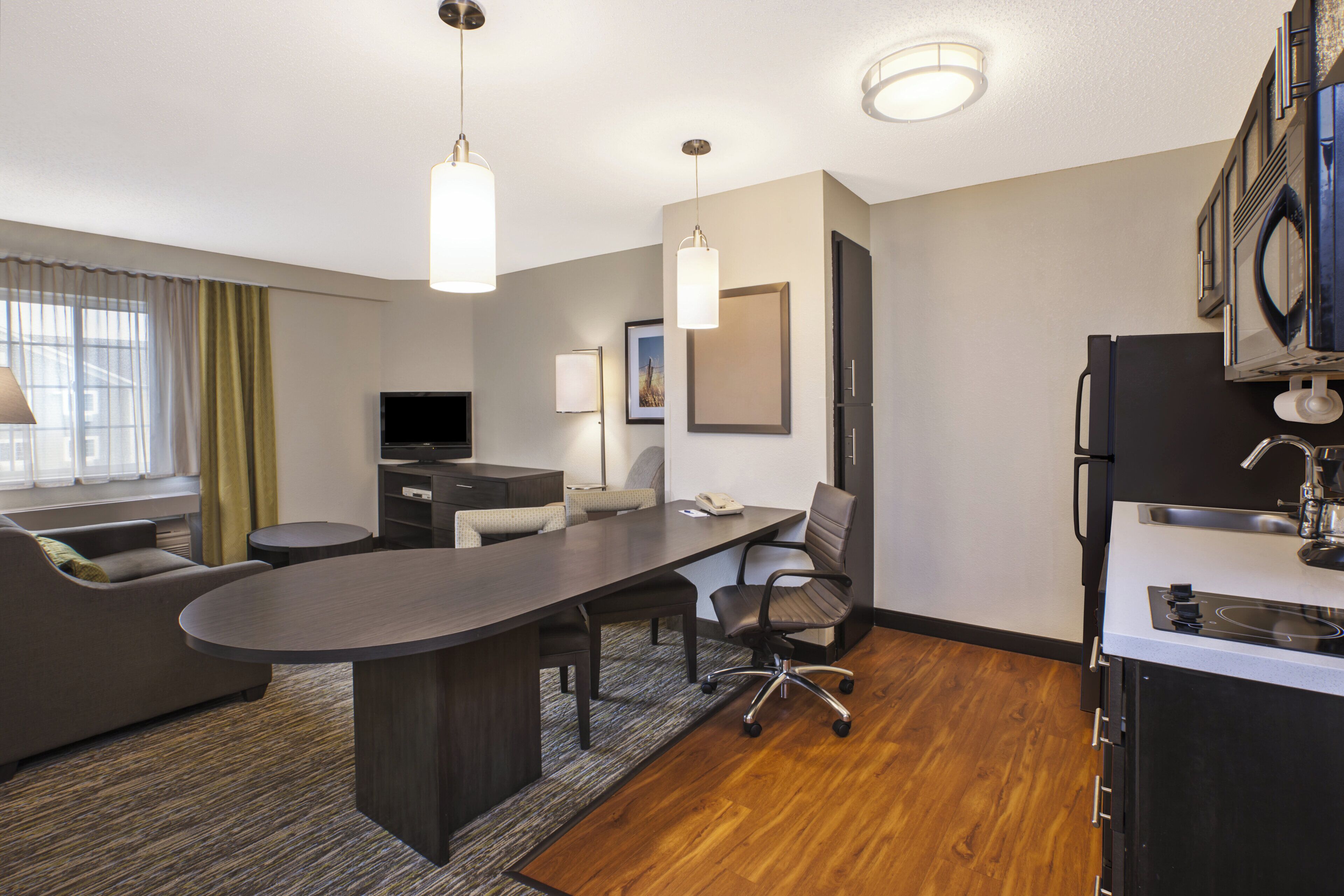 A hotel room with a kitchenette, a seating area with a sofa and chairs, a flat-screen TV, and a large window with curtains.