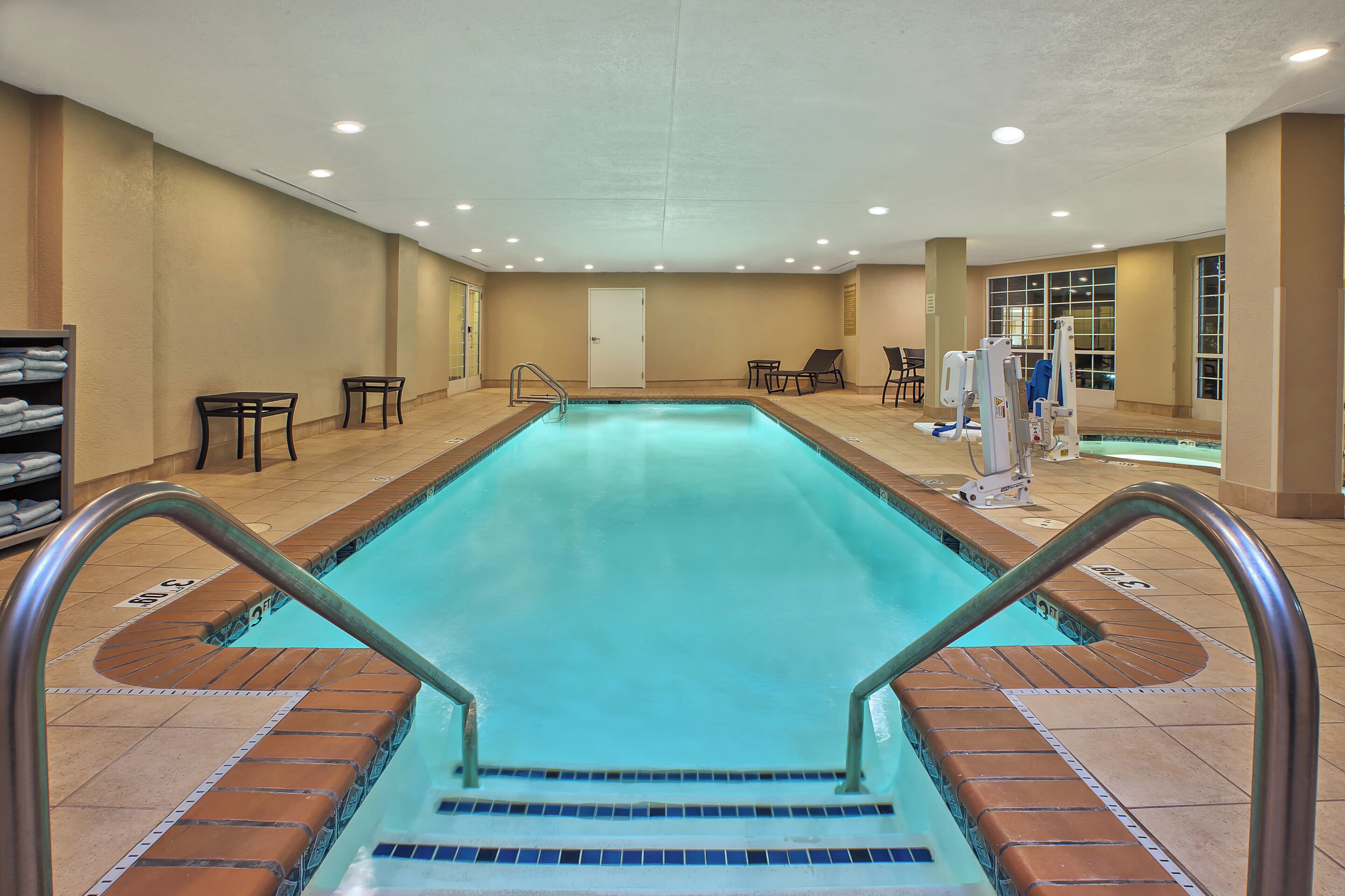 An indoor swimming pool with a clear water surface, surrounded by a tiled deck, and equipped with a lifeguard stand and lounge chairs.