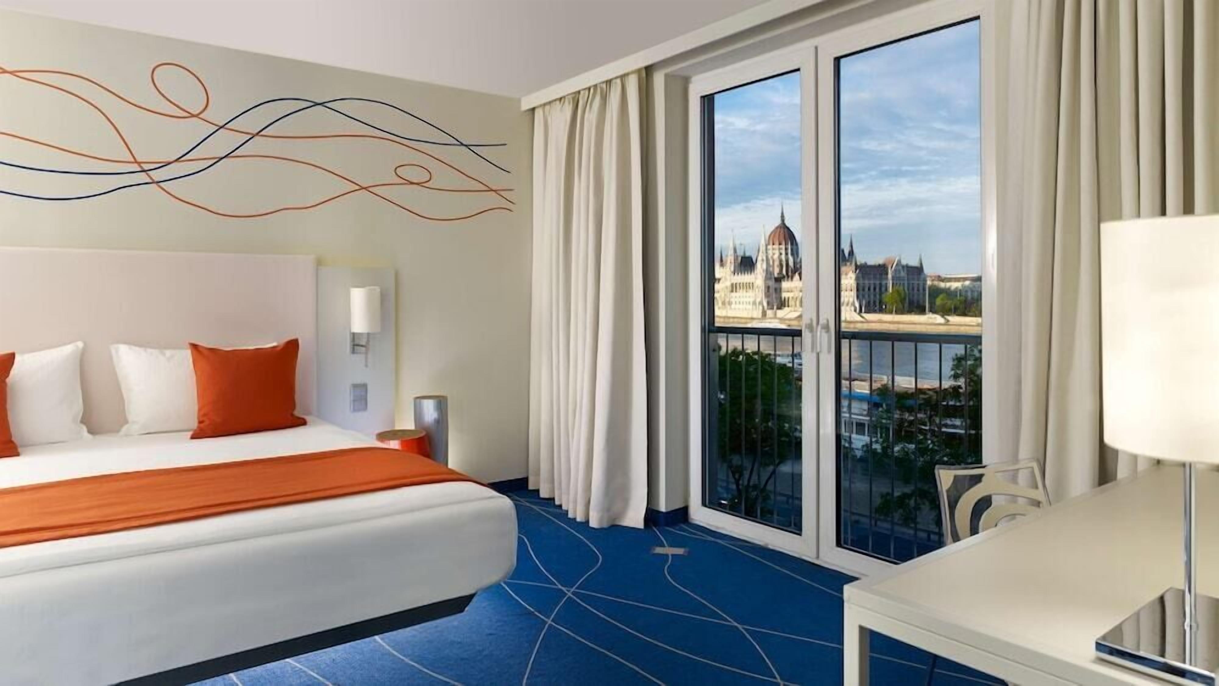 Habitació d'hotel amb un llit gran, un escriptori i vista a un edifici històric a través d'una porta de vidre.
