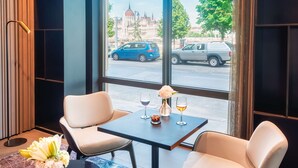 Ein modernes Hotelzimmer mit großem Fenster und Blick auf die Stadtlandschaft, darunter ein markantes Gebäude. Ein kleiner Tisch mit zwei Stühlen und eine Vase mit Blumen stehen ebenfalls im Raum.