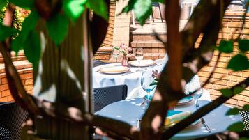 Un área de comedor al aire libre con mesas preparadas para dos personas, rodeada de muros de ladrillo y follaje.