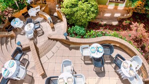 Eine Außenpatio-Terrasse mit mehreren Tischen und Stühlen, umgeben von üppiger Bepflanzung und einer Zierwand mit Fenstern.