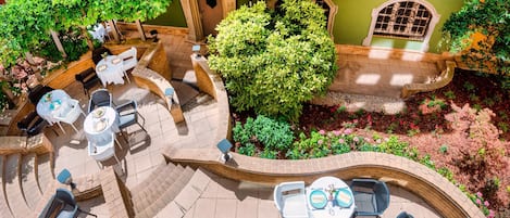 An outdoor patio area with multiple tables and chairs, surrounded by lush greenery and a decorative wall with windows.