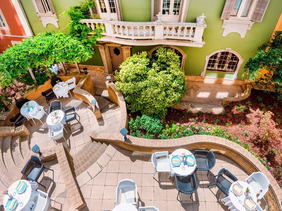 Un patio al aire libre con varias mesas y sillas, rodeado de abundante vegetación y un muro decorativo con ventanas.
