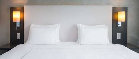 Cama doble con sábanas blancas, dos mesitas de noche con lámparas y una pared texturizada detrás de la cama.